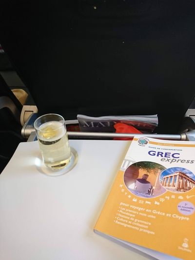 Guide de conversation Grec Express et verre de Champagne posés sur la tablette d'un siège d'avion en plein vol vers Athènes.