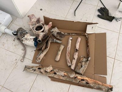 The image shows various pieces of heavily corroded and damaged plumbing pipes and fittings laid out on a piece of cardboard on a tiled floor.